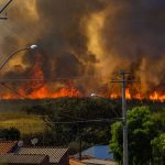 Incêndio florestal se espalha próximo a casas, com chamas e fumaça intensa.