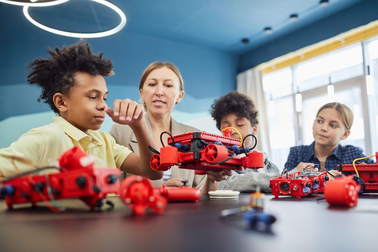 Estudantes e uma professora colaboram na construção de um projeto de robótica em uma sala de aula.